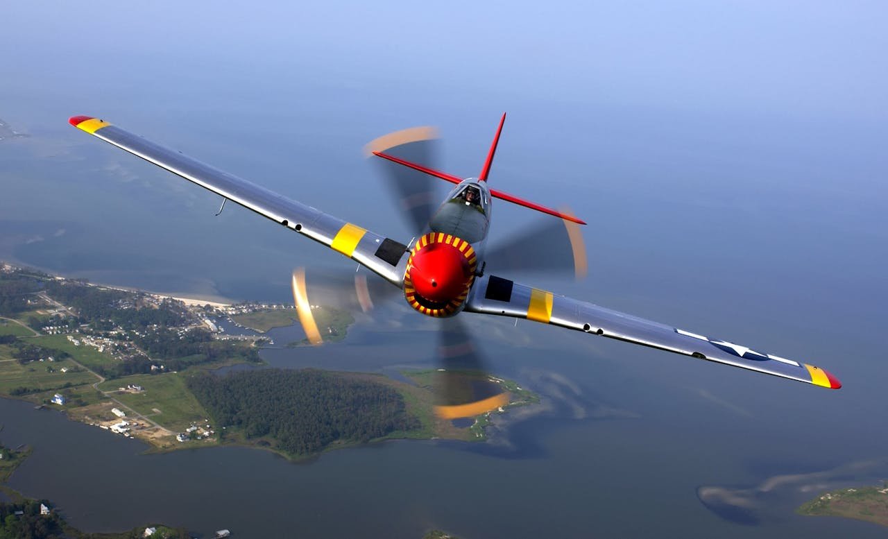 A vintage fighter aircraft, P-51 Mustang, flying over coastal landscape with propeller motion blur.